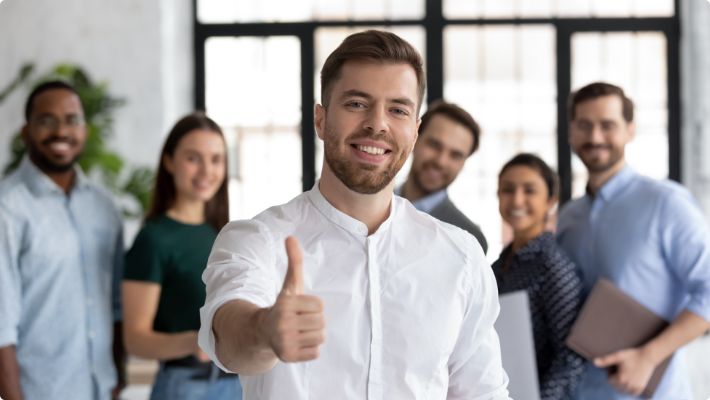 Mit der richtigen Umschulung in eine sichere berufliche Zukunft - zu sehen ist ein junger man im Vordergrund, der lächelnd in die Kamera schaut und einen Daumen hoch zeigt. Im Hintergrund ist das Team zu sehen.